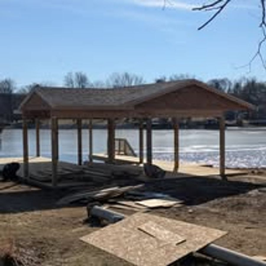 Waterfront pavilion under construction with post-and-beam roof framing beside a lake